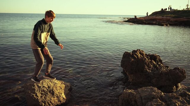 Young adventurous boy happily jumping from one rock to another over sea during beautiful sunset, embody sense of freedom, youth, and enjoy outdoor family activities