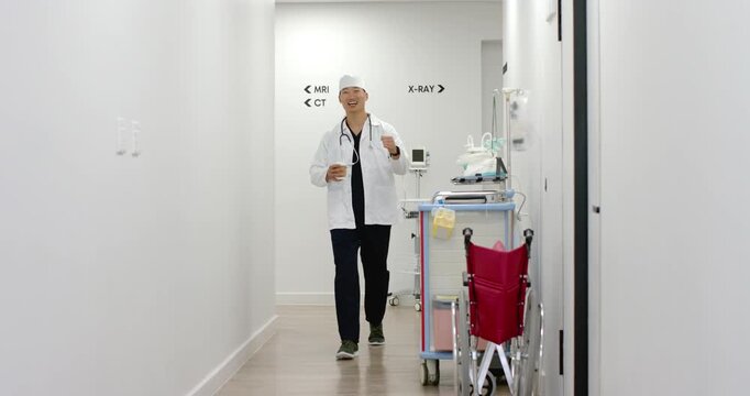 Asian man walking hall to camera MRI or CT or XRAY signs with stethoscope holding cup smiling laughi