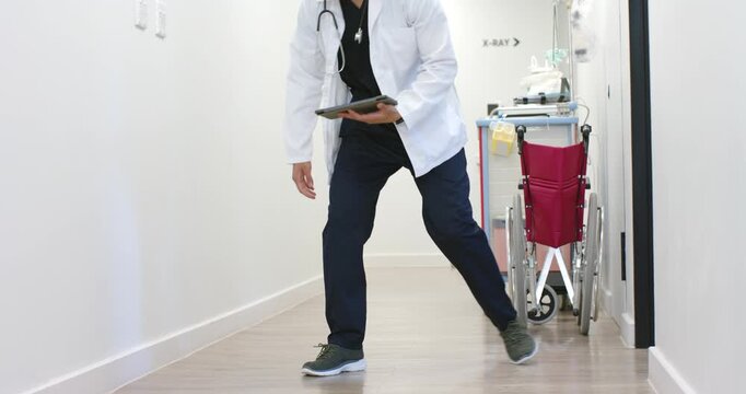 Male MD in lab coat walking in hospital viewing tablet pausing, sitting by red wheelchair, cart