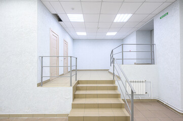 Obraz premium clean, minimalist hallway with beige tiled stairs, metal railings, white textured walls, recessed ceiling lights, wooden doors, radiator, and green «EXIT» sign. Functional, uncluttered design.