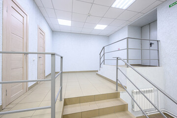 Obraz premium clean, minimalist hallway with beige tiled floor, white textured walls, metal railings, wooden doors, staircase, recessed ceiling lights, and white radiator. Functional, uncluttered space.