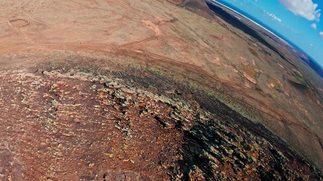 Aerial view of Fuerteventura's volcanic interior shows crater rims, lava slopes, sparse scrub, Atlantic horizon, dirt tracks, hikers, and midday clouds.