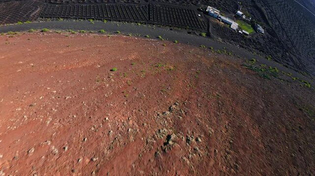 Aerial drone shot glides over La Geria pits and stone windbreaks in Lanzarote, with black ash, orange brown vine stumps, semicircular walls, and distant volcanic ridges.