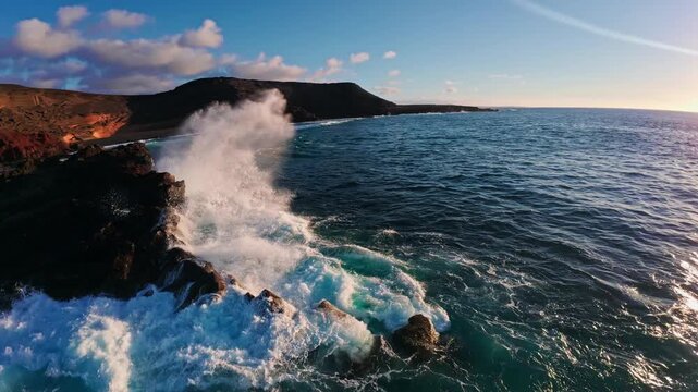 Aerial camera tracks low along El Golfo and Los Hervideros in Lanzarote as turquoise waves hit dark basalt and rust red cliffs in warm golden hour light