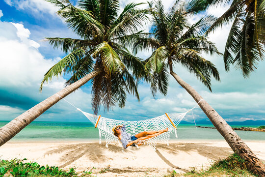 Happy beautiful girl in a straw hat and shorts, striped t-shirt, lying on a beach hammock between two palm trees, on the seashore of a tropical island, traveling summer vacation