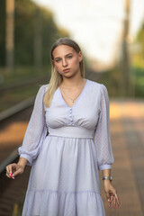 Fototapeta premium Young woman with long blonde hair wearing a light purple dress stands on a train platform, soft sunlight illuminating her figure and the railway tracks in the background