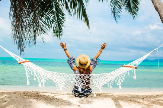 Happy beautiful girl in a straw hat and shorts, striped t-shirt, lying on a beach hammock between two palm trees, on the seashore of a tropical island, traveling summer vacation