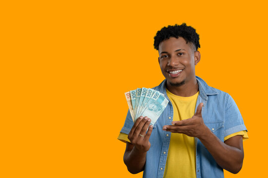 A happy young man holding Brazilian Real banknotes and smiling. Orange background.