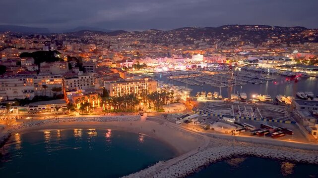 Aerial Footage Showcases Vibrant Waterfront Illumination Against City And Mountain Skyline Backdrop. Cannes. France
