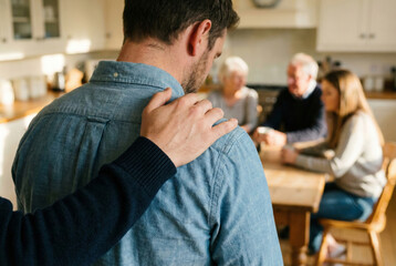 A supportive hand rests on the shoulder of a man in a denim shirt, viewed from behind, while a concerned family group, including an elderly couple, sits at a kitchen table in the blurred background.