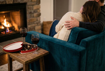 A rear view of a romantic couple embracing on a teal velvet armchair in front of a warm fireplace, accompanied by glasses of red wine and a heart-shaped box of chocolates on a wooden side table.