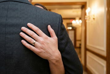 A close-up detail shot of a hand wearing a silver wedding band gently resting on the back of a man in a dark grey formal suit, set against a blurred background of a luxury hallway with a chandelier.