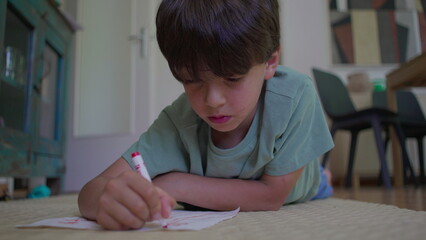 Boy draws on paper while resting on elbows, lying on floor at home, immersed in quiet creative activity using red marker