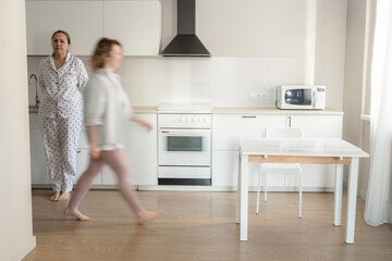 A grown daughter rushes past her mother in bright, modern kitchen. A busy family morning in minimalist white kitchen, motion blur