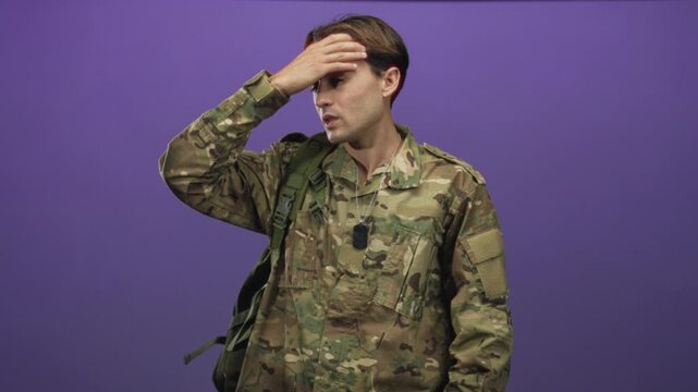 Man in camouflage uniform touches forehead with hand, dog tags and backpack visible in studio; exhaustion resilience.