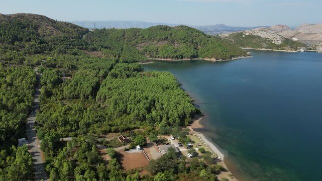 Aerial drone view of a small campsite and RV parking area on the forested shores of Lake Komani, Albania, featuring turquoise water and rugged mountain peaks in the Balkan wilderness.