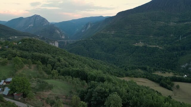 Aerial view of a rafting campsite nestled in the lush Tara River Canyon, Montenegro, with the historic Durdevica Tara Bridge visible in the distance between the steep, forested mountain peaks.