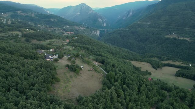 Aerial view of a rafting campsite nestled in the lush Tara River Canyon, Montenegro, with the historic Durdevica Tara Bridge visible in the distance between the steep, forested mountain peaks.