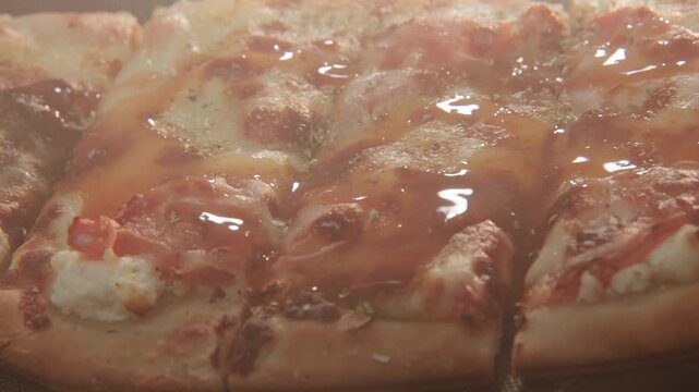 Red juicy hot sauce slowly in slow motion drips and pours onto square cut tasty pizza from pizzeria restaurant takeout as juices, herbs, and sauces spill over cheese, crust, and bread on surface