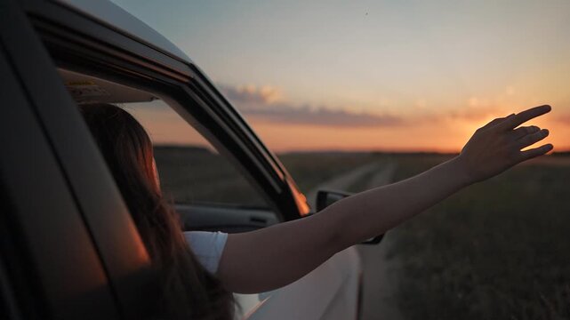 Open hand of girl outside car window catches air. Summer sunset lights up road. Freedom begins with each hand motion. Girl leans from car toward sunset. Road becomes symbol of hand and freedom.