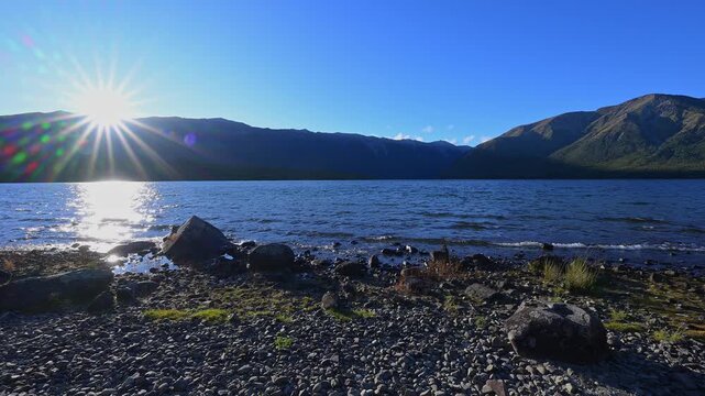Ein ruhiger See mit Sonnenstrahlen, die auf das Wasser und Ufer treffen, umgeben von bewaldeten Bergen unter klarem Himmel, Sommer, Lake Rotoiti, Saint Arnaud, Tasman Region, S&uuml;dinsel, Neuseeland, Oze