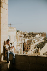 Woman sitting on wall in Malta with city view while using phone