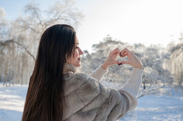 Woman in fur coat forming heart with hands in snowy park, winter emotional concept