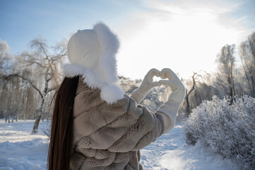 Woman in fur coat making heart shape with hands in snowy park, winter love concept
