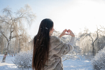 Woman in fur coat forming heart with hands in snowy park, winter emotional concept