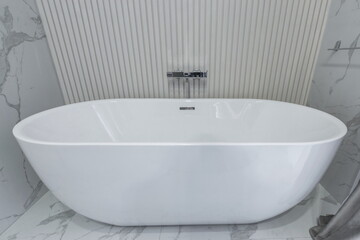 A white bathtub is shown in a bathroom with light marble tiles on the floor and a textured wall behind it. The faucet and taps are chrome with a modern design