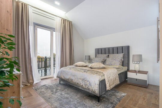 This bedroom features a bed with soft bedding and pillows. Large windows bring in natural light, and curtains add warmth. A small table and a green plant complete the space
