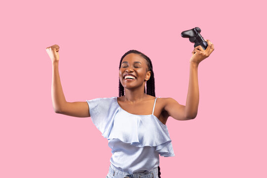 Excited female gamer with joystick making YES gesture, feeling overjoyed over her victory, pink studio background. Young black woman with controller playing computer games