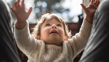 Happy toddler with curly brown hair reaches up with both hands while looking up with joy, wearing a cozy knitted sweater in a backlit scene with a soft focus background