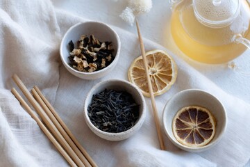 Top view of herbal tea preparation with glass teapot and dried ingredients. Bamboo straws and ceramic bowls on white linen cloth. Healthy organic lifestyle concept