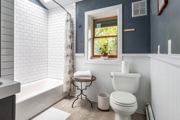 Bright bathroom interior with white subway tile bathtub, gray walls, toilet and window with natural...