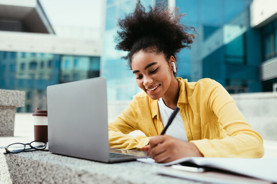 Smiling Black Student Girl At Laptop Taking Notes Learning Online And Doing Homework Sitting At Modern University Building Campus Outdoor. Distance Education, E-Learning Concept