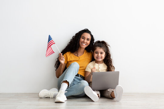Learn Online. Little Girl And Her Mom Holding Laptop And American Flag While Sitting On Floor Near White Wall At Home, Mother And Little Daughter Study Foreign Languages, Enjoying Remote Education
