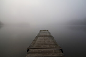 Obraz premium A foggy morning on the lake creates an atmosphere of peace and mysticism. A wooden pier, extending into the thick fog, emphasizes the depth and infinity of space. A minimalist Finnish autumn landscape