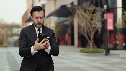Portrait of elderly company manager navigates with smartphone as walks across downtown block. Experienced entrepreneur in suit holds mobile phone planning route in urban district