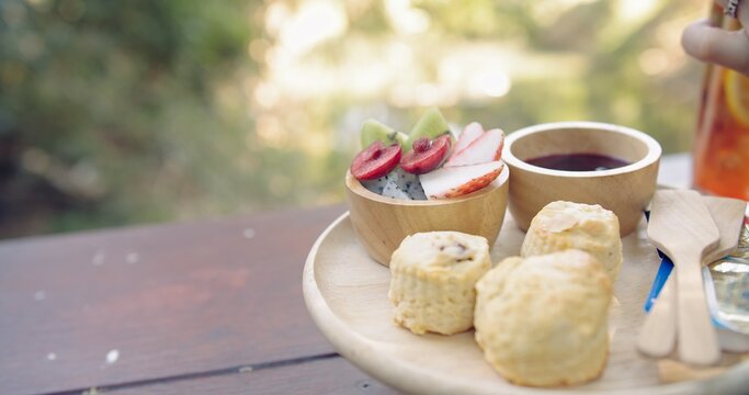 Delicious scones and fresh fruit platter served on a wooden tray with jam and drinks in a serene outdoor garden setting, evoking a relaxing and wholesome brunch experience.