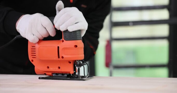 Worker in gloves holds electrical jigsaw cutting wooden board slowly. Power tool vibrates on surface while shaping straight line on panel in workshop