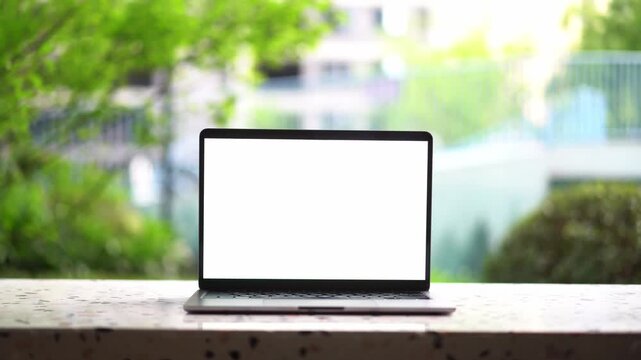 Laptop with blank white screen placed on wooden windowsill against blurred green plant background