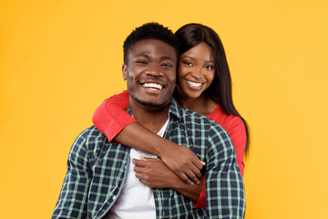 Fototapeta premium A man and a woman embrace each other while smiling. They stand in front of a bright yellow background. The man wears a checkered shirt, and the woman wears a red outfit.