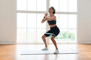 Fototapeta premium Training Concept. Confident determined sportswoman squatting with elastic resistance band in fitness studio or at home in living room doing leg and buttocks workout, listening to music in earbuds