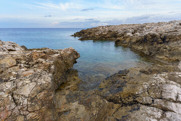A wide, scenic view of the rugged, textured limestone coastline of Cala Minnola, Levanzo Island, meeting the vast, deep blue Mediterranean Sea under a dramatic, cloud-streaked sky.