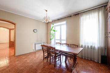 Fototapeta premium Spacious dining room with wooden table and chairs, chandelier, sheer curtains, large window, terracotta tiled floor, wall clock, radiator, arched doorway, potted plant. Light, airy ambiance.