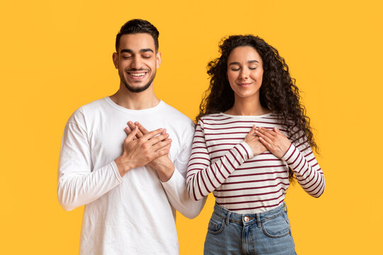 Thank You. Grateful Young Arab Man And Woman With Eyes Closed Keeping Both Hands On Chest, Thankful Middle Eastern Couple Expressing Grattitude And Kindness While Posing Over Yellow Background