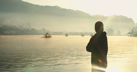 A solitary individual stands contemplating the serene misty lake at sunrise with traditional boats and distant mountains, embodying peace and journey in a beautiful landscape. © HarryKiiM Stock