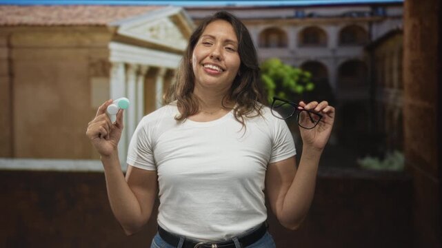 Woman holds glasses and contact lens case at chest level in old town building courtyard, eyes closed and smiling with relaxed posture; relief ease.