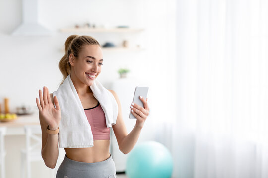 Online call and fitness blog, modern tech for chat, body care and workout with coach. Glad millennial blonde female in sportswear with towel waving hand to smartphone webcam, in living room interior - Powered by Adobe
